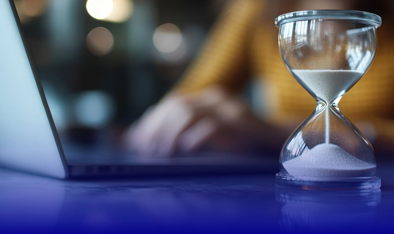 Blog | Image of hourglass sitting on a desk with a person working on a laptop